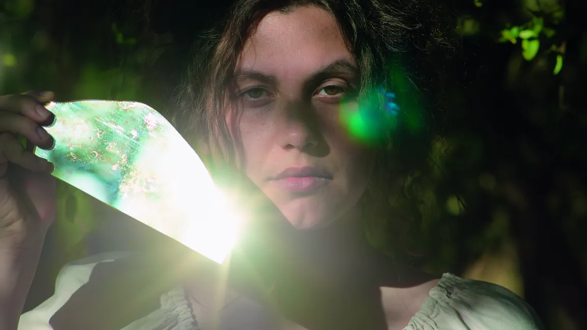 Filmstill Junge Frau mit dunklen Haaren die einen Spiegel neben ihrem Gesicht hält, der die Sonne reflektiert