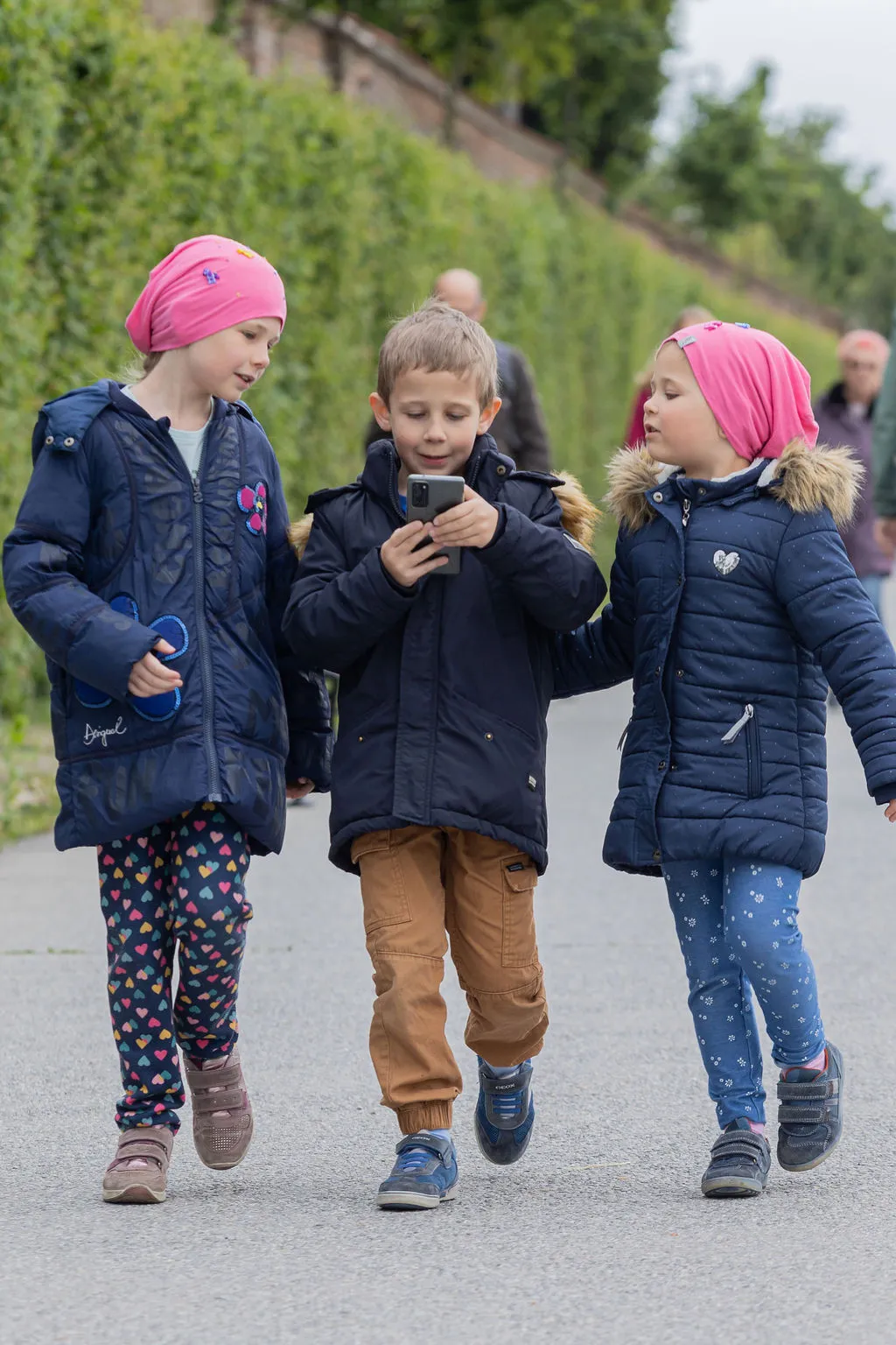 Drei Kinder - zwei Mädchen mit rosa Hauben und ein Bub in der Mitte, laufen durch die Gartenanlage und spielen am Handy ein VR-Spiel.