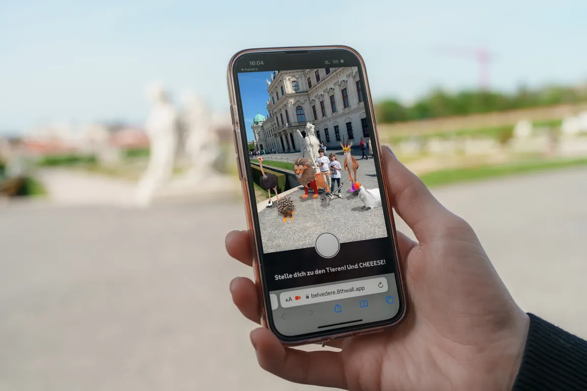 Ein Smartphone-Screen mit einer Ansicht des Fantastic Palastics: Garden Game. Das Handy wird in der Gartenanlage hochgehalten - im Hintergrund sieht man den Schlosspark.