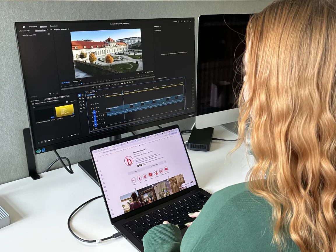 Zwei Computermonitor, auf einem wird ein Video geschnitten, auf dem anderen ist eine Website geöffnet. Im Vordergrund ist eine Person mit langen roten Haaren von hinten angeschnitten sichtbar.