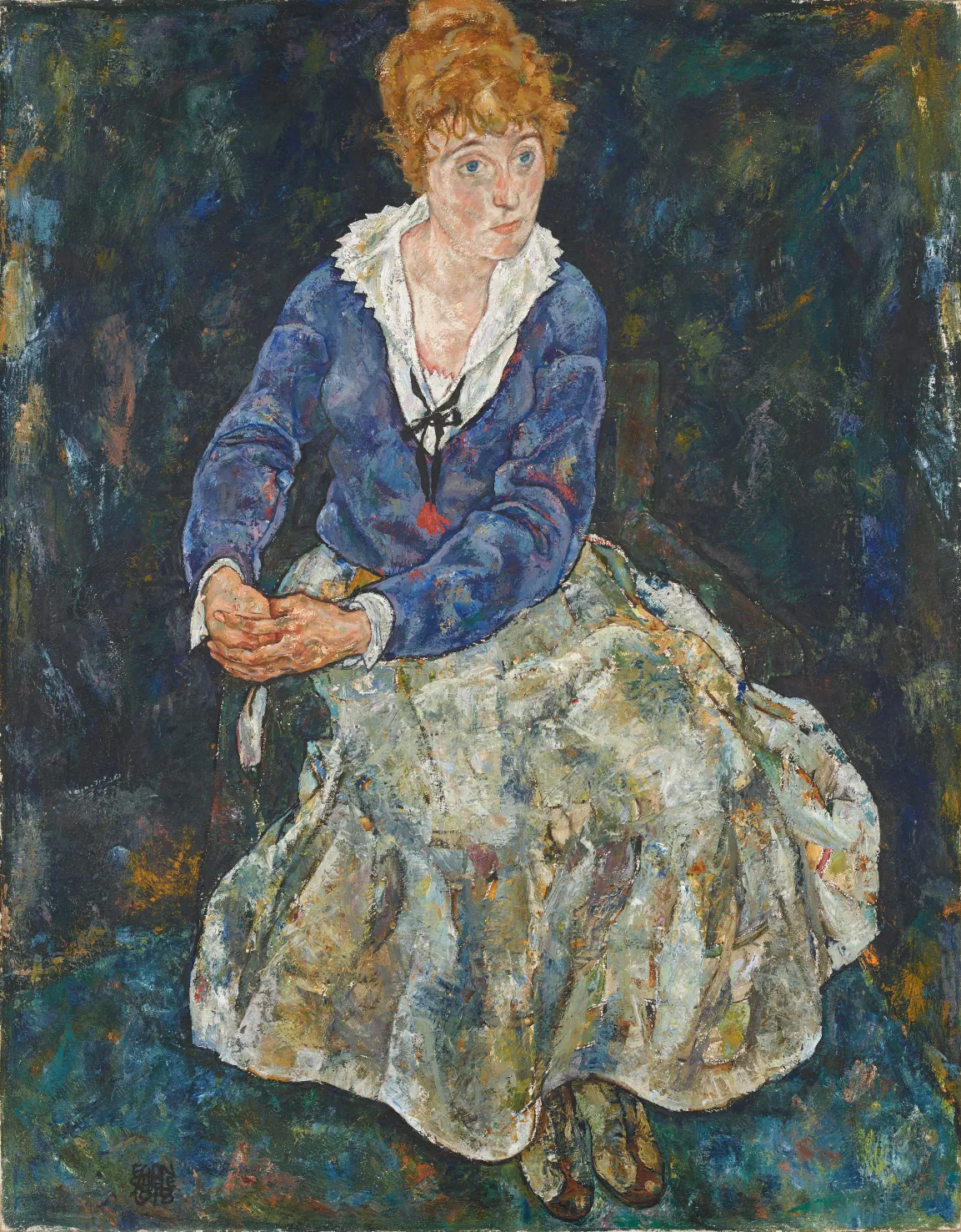 A seated woman wearing a white blouse and blue sweater over a cream colored skirt. She wears her dark blonde hair in a bun.