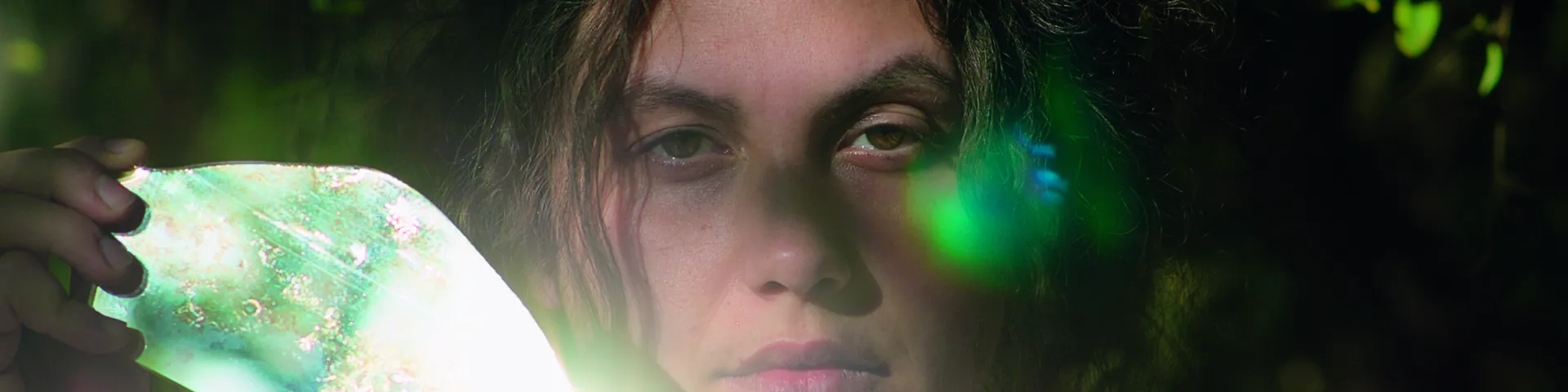 Filmstill Junge Frau mit dunklen Haaren die einen Spiegel neben ihrem Gesicht hält, der die Sonne reflektiert
