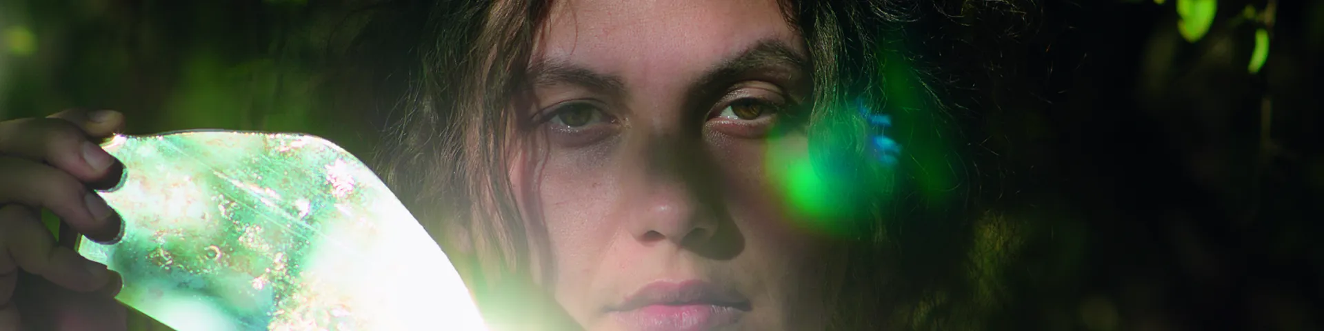 Filmstill Junge Frau mit dunklen Haaren die einen Spiegel neben ihrem Gesicht hält, der die Sonne reflektiert