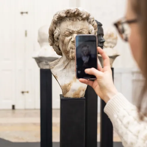 Eine Frau macht mit dem Handy Fotos von einem Charakterkopf von Franz Xaver Messerschmidt im Ausstellungsraum