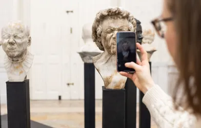 Eine Frau macht mit dem Handy Fotos von einem Charakterkopf von Franz Xaver Messerschmidt im Ausstellungsraum